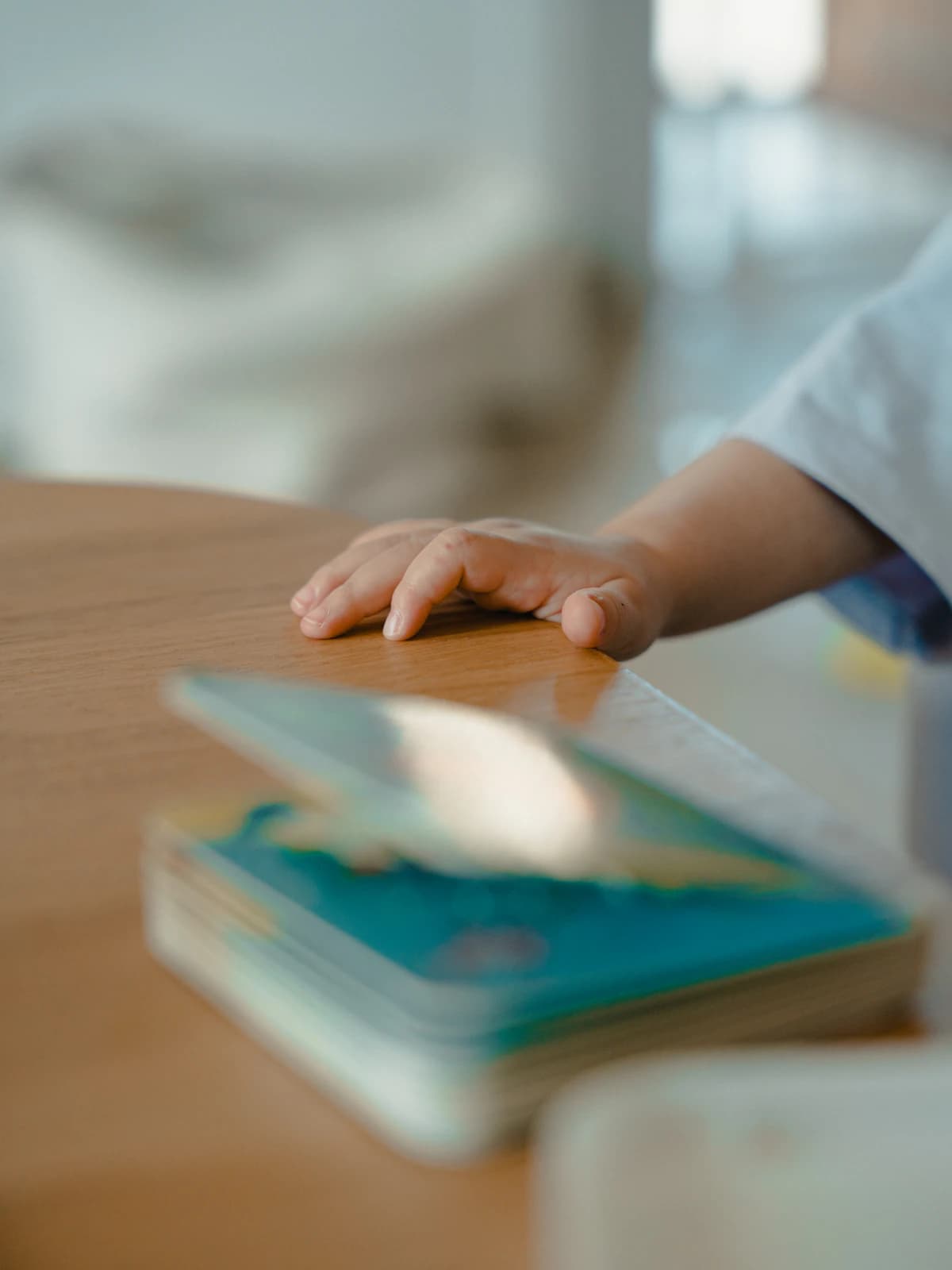A toddler's hand reaching for a picture book on a table in soft daylight